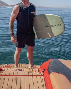 Lifestyle image of man holding the Keowee wake surf board in green, while wearing a black front zip quilted pattern competition impact wake surf vest with black shorts. 