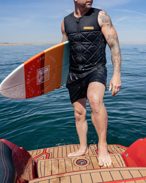 Lifestyle image of man holding the Ozark wake surf board with retro colors while wearing a black front zip quilted pattern competition impact wake surf vest with black shorts. Man is standing on the back of a G23 Nautique. 