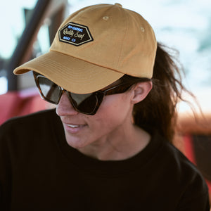 Lifestyle image of woman driving boat with dad hat from Pit and Barrel Surf co in camel color. 