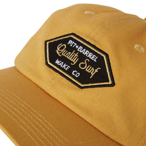 Detail view of gold dad hat that is canvas with 'Pit + Barrel Quality Surf Wake Co' patch in black with white and gold font. 