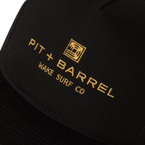 Detail view of black 5 panel snapback cap with gold text 'pit + barrel wake surf co' and curling wave logo on a white background