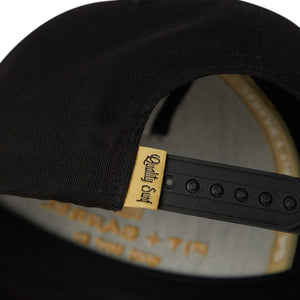 Detail view of black snapback 5 panel hat with black closure and 'quality surf' text tag in gold with black letters. 