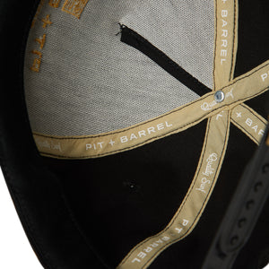 interior detail view of black 5 panel snapback. Seam tape with 'pit + barrel' and 'quality surf' text in gold and white lettering. 