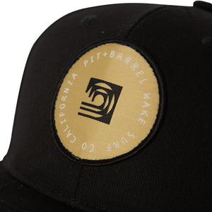 Detail view of black 5 panel snapback with gold logo patch that includes a black curling wave and white 'Pit + Barrel wake surf co California' 