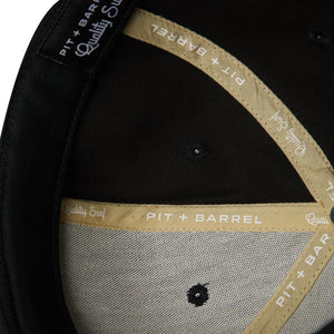 Detail view of interior of  black snapback hat with gold seam tape with white lettering that says 'Pit + Barrel Quality Surf'. 
