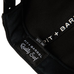 detail view of black snapback cap with focus on 'Pit + Barrel Quality Surf' hidden tag. 