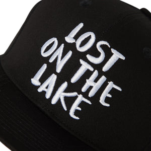 Detail view of a black 5 panel snapback cap with 'Lost on the Lake' text in white on a white background.