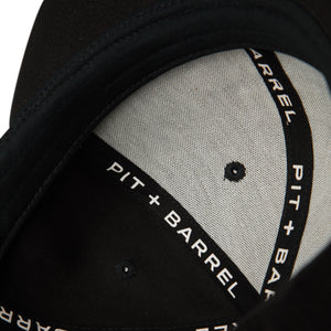 Interior detail view of black 5 panel snapback with black seam tape with text saying 'Pit +Barrel' in white font. 