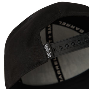 detail view of black snapback with tag on closure that is black with white lettering that says ' Quality Surf'. 