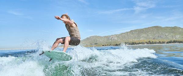 Lifestyle image of pro wake surf rider in Pit and Barrel Tan Slasher vest riding the Keowee wake surf board doing an air behind a boat on a lake. 